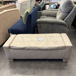 A beige upholstered bench sits in a furniture store among sofas and chairs on a concrete floor.