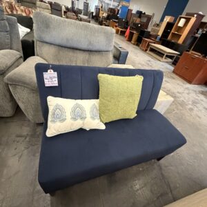 A blue loveseat with two decorative pillows, displayed in a furniture store with other sofas and shelves.