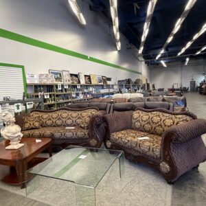 Two ornate brown sofas and glass tables in a large thrift store with shelves of books and decor in the background.