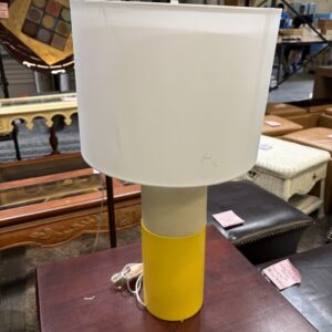 A modern table lamp with a yellow base and white shade sits on a wooden table in a store.