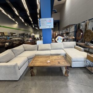 Large beige sectional sofa with a wooden coffee table in a furniture store, blue pillar in the background.