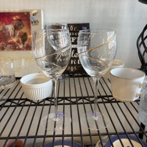 Two empty wine glasses with gold accents on a metal shelf, surrounded by various cups and dishes.