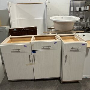 Three empty white kitchen cabinets without tops are lined up in a store, with a sink and other items nearby.