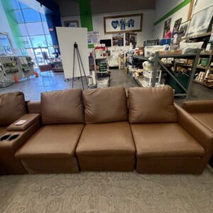 Brown leather sofa with cup holders in a store with various items and a display board in the background.