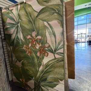 Large area rug with green tropical leaves and pink flowers displayed in a store near a window.