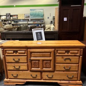 A wooden dresser with multiple drawers sits in a store with a framed sign and furniture in the background.