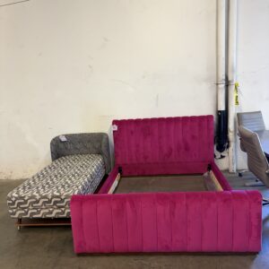 A pink velvet bed frame and a patterned chaise lounge sit in a warehouse-like space with a plain wall.