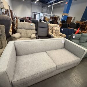 A light gray sofa on display in a furniture store, surrounded by various chairs and tables.