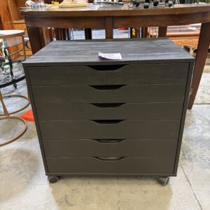Black six-drawer rolling cabinet with curved handles, placed on a concrete floor in a furniture store.