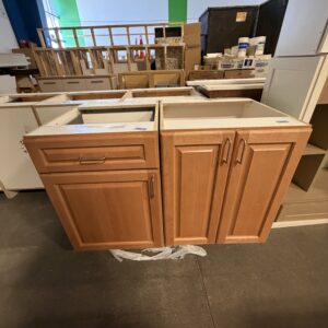 Wooden kitchen cabinets on display in a store, placed on a dark floor with various items in the background.
