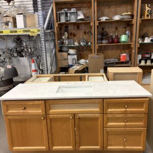 A wooden kitchen island with drawers and a marble countertop sits in a store with shelves of various items behind it.