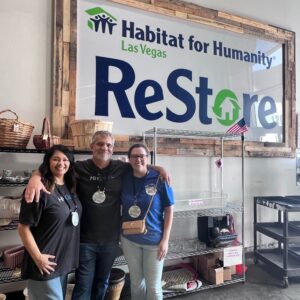 Three people smile and pose together inside a Habitat for Humanity Las Vegas ReStore, standing by empty shelves.