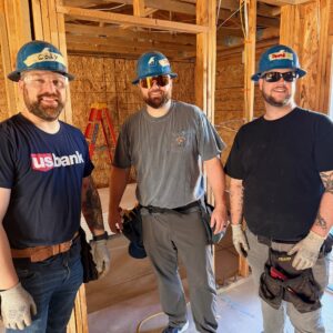 Three men in hard hats and work gloves stand in a partially constructed wooden building, smiling at the camera.