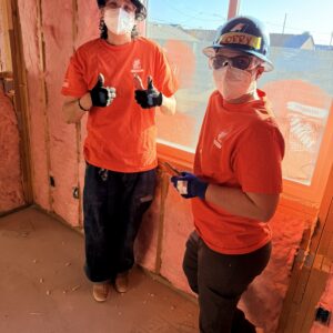 Two people in orange shirts and hard hats give thumbs up inside a building under construction.