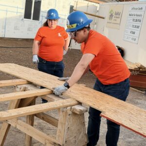 Two people in blue hard hats and orange shirts cut wood at a construction site with a Habitat for Humanity sign.