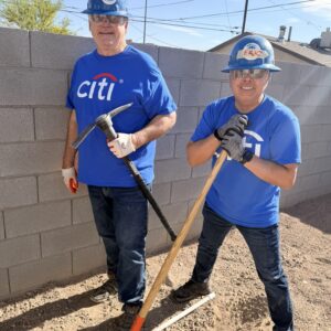 Two men in blue Citi shirts and hard hats stand outdoors with shovels, working near a brick wall.