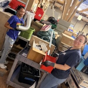 Two women organize and sort supplies in a cluttered storage room filled with boxes and various items.
