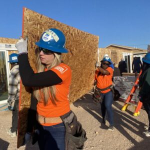 Two women in hard hats and orange shirts carry a sheet of plywood at a construction site on a sunny day.
