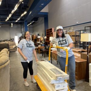 Two women in HGV shirts move boxes on a yellow cart in a furniture-filled warehouse, smiling at the camera.