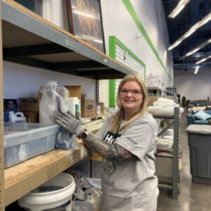A woman in a gray shirt organizes shelves in a warehouse, smiling at the camera.