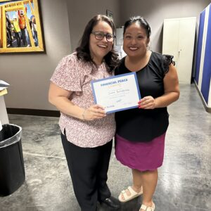 Two women smile as one holds a Financial Peace certificate in an office with art on the wall.