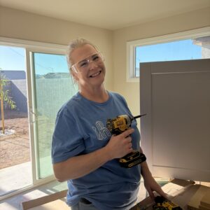 Smiling woman in safety glasses holding a power drill, assembling furniture in a sunlit room.