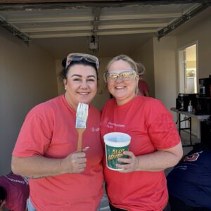 Two women in red shirts smiling, one holding a paintbrush, the other a cup, standing in a garage.