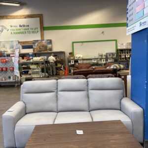 Light gray sofa and wooden coffee table in a thrift store with shelves, signs, and a person in the background.