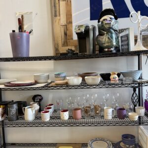 Shelves displaying assorted kitchenware, mugs, glasses, plates, bowls, and a coffee maker in a shop.