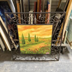 Framed landscape painting with trees and flowers, displayed among stacked artworks in a warehouse or storage area.