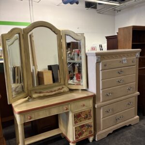 A beige vanity with a tri-fold mirror and a matching chest of drawers displayed in a thrift store.