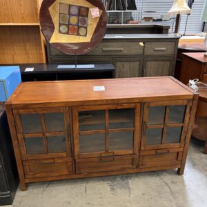 A wooden cabinet with glass doors and drawers is displayed in a store with other furniture and decor items.
