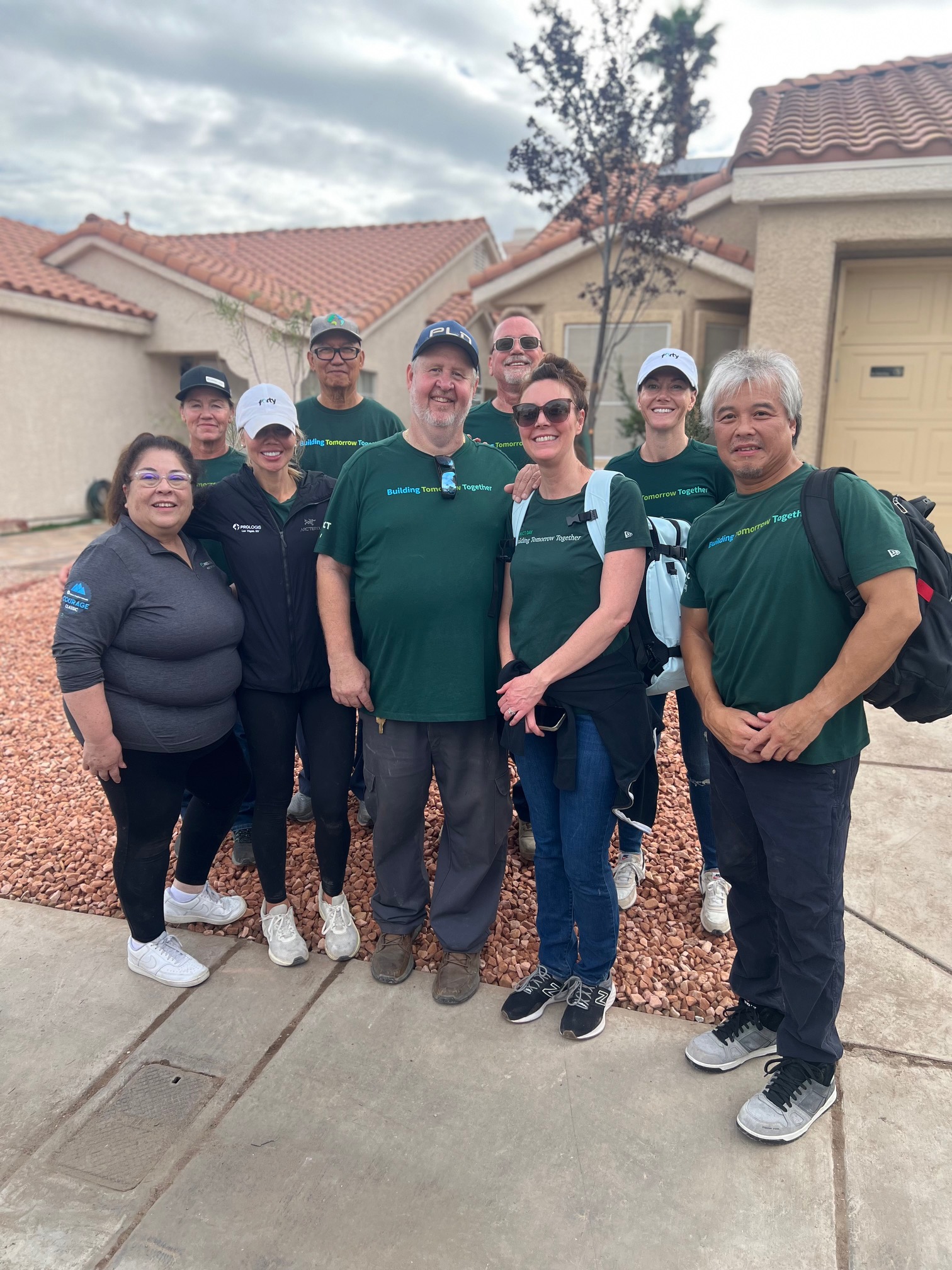 Group of smiling people in green shirts posing outside a house with rocks in the yard on a cloudy day.