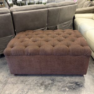 A brown tufted ottoman sits on a concrete floor in a furniture store with sofas and paint cans in the background.
