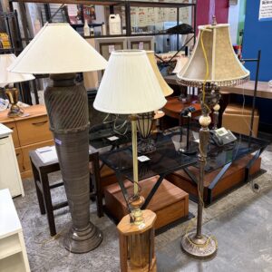 Three floor lamps with lampshades displayed in a thrift store among wooden tables and other furniture.