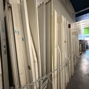 Various doors and panels stand upright in a row behind a chain inside a store or warehouse.