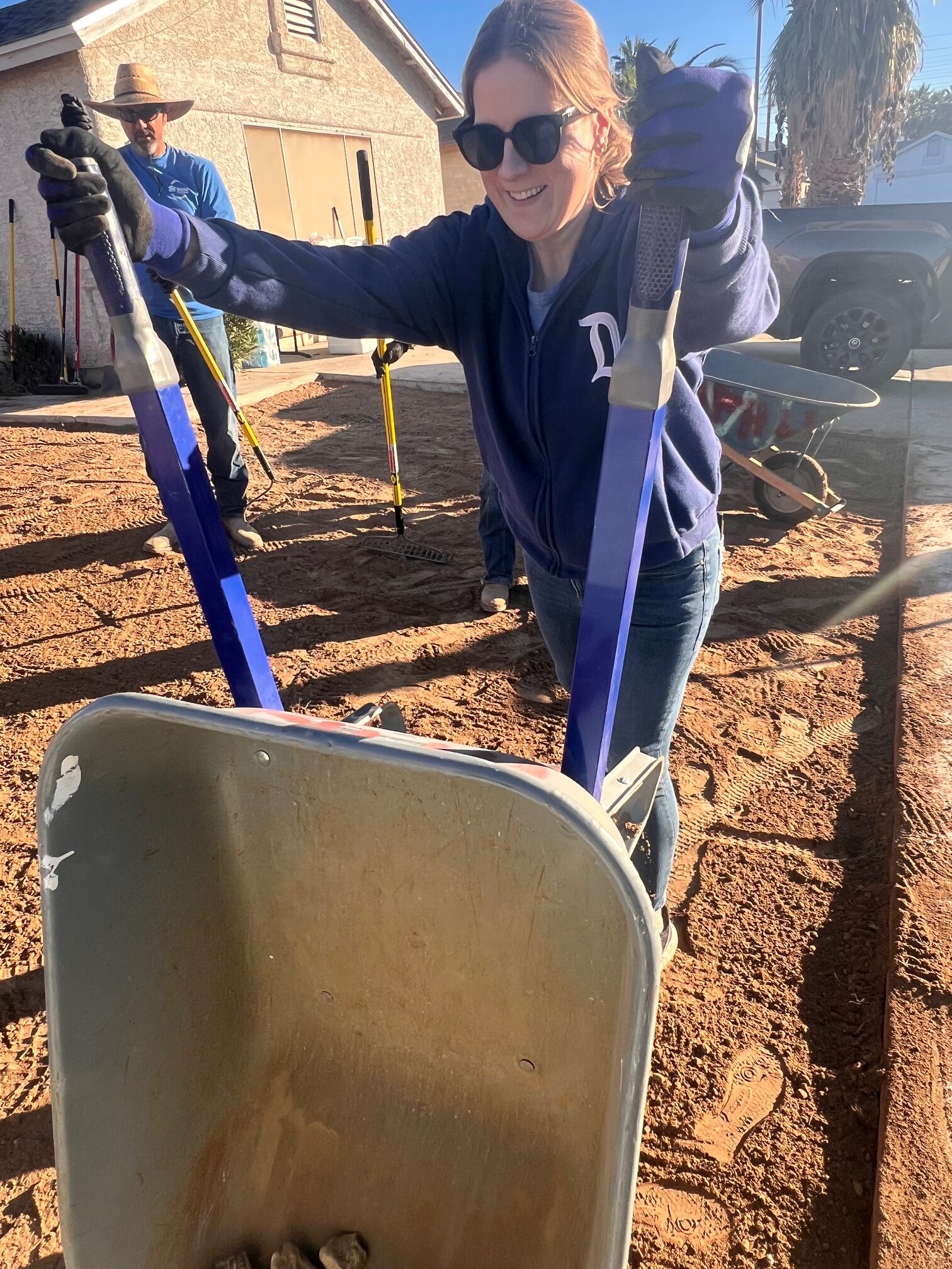 Woman in sunglasses using a wheelbarrow on a sunny day, with a man and yard tools in the background.