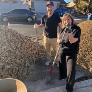 Two people with shovels stand by piles of rocks on a sunny day in a residential neighborhood.