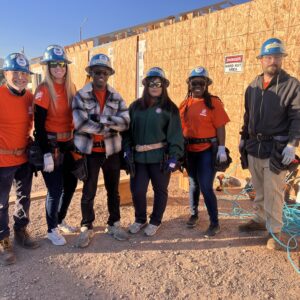 Six construction workers in hard hats and safety gear pose together at a building site in the sunlight.