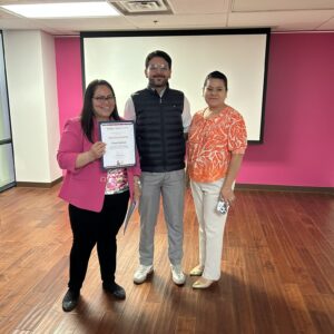 Three people stand indoors; one woman holds a certificate and smiles, in front of a pink and white wall.