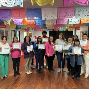 A group of people stand indoors holding certificates under colorful papel picado decorations.