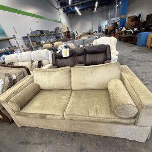 Tan sofa with two round pillows displayed in a furniture store, surrounded by other sofas and shelves.