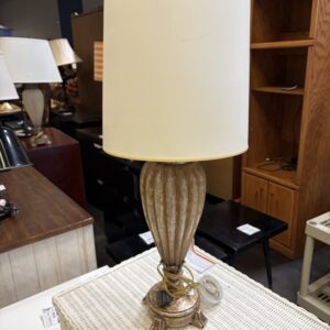 Beige lamp with pleated base and round shade sits on a white wicker table in a furniture store.