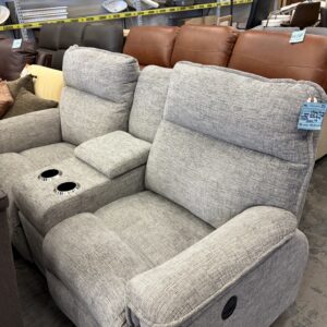 Gray reclining loveseat with cup holders on display in a furniture store surrounded by other recliners.