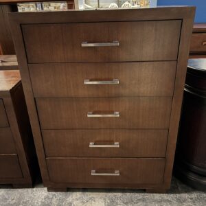 A tall wooden dresser with five drawers and silver handles, standing on a concrete floor in a store.