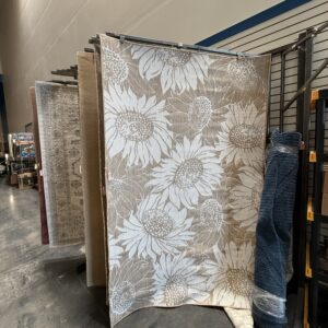 Large beige rug with white sunflower pattern displayed upright in a store aisle among other carpets.