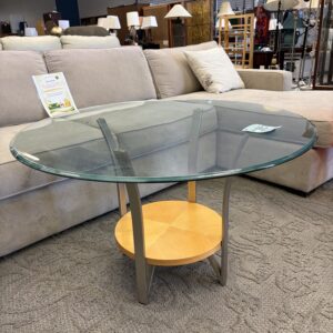 Round glass coffee table with metal legs and a light wood shelf, placed in front of a beige sofa.
