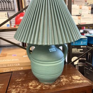 A teal ceramic table lamp with a pleated shade sits on a brown wooden surface in a store.