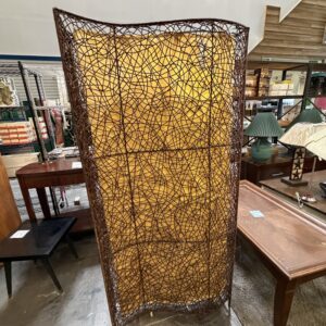 A brown woven room divider with a yellowish background stands in a store among wooden tables and lamps.