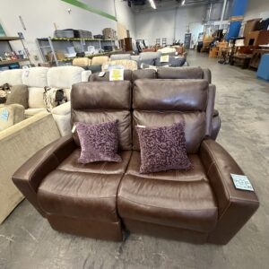 Brown leather loveseat with two purple accent pillows in a furniture store; shelves and sofas in background.